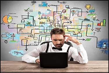 Man looking stressed at a laptop with complex logistics diagrams on the wall